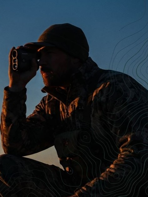 Hunter using a rangefinder at sunset, wearing camouflage and a beanie, focused on spotting wildlife in the field.